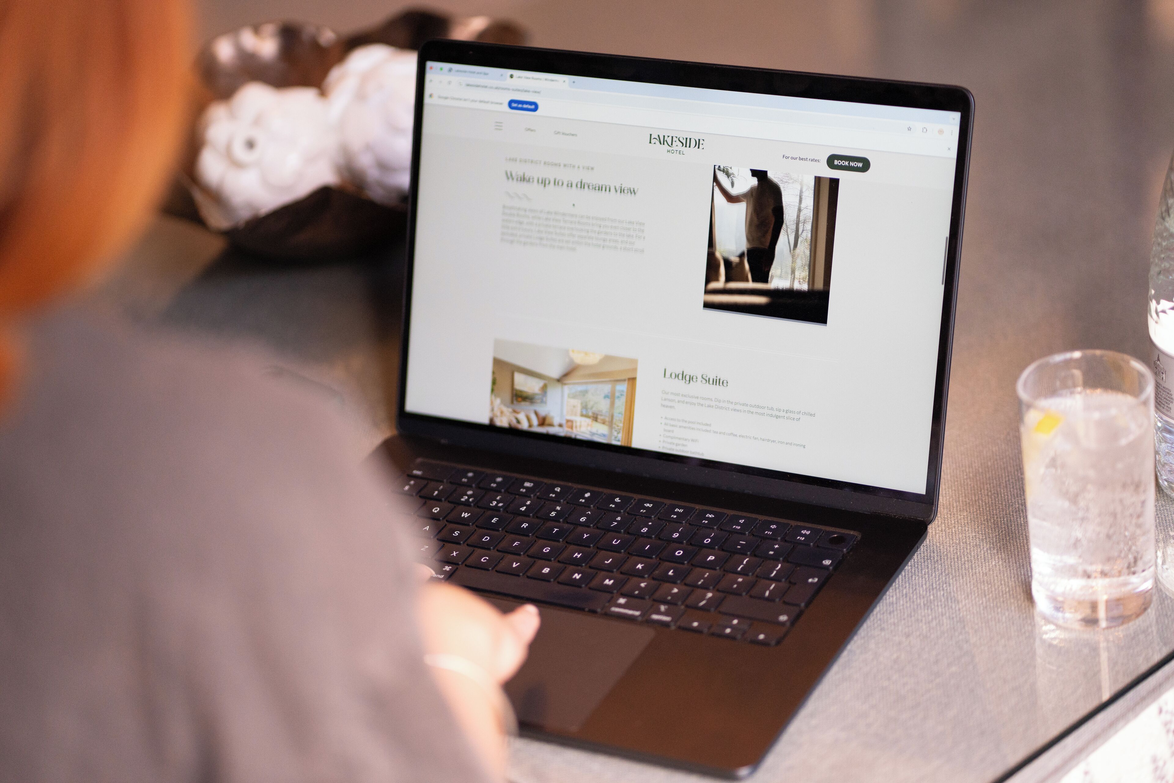 Una laptop muestra un sitio web de hotel con una sección titulada 'Lodge Suite' y una foto de una persona de pie junto a una ventana.