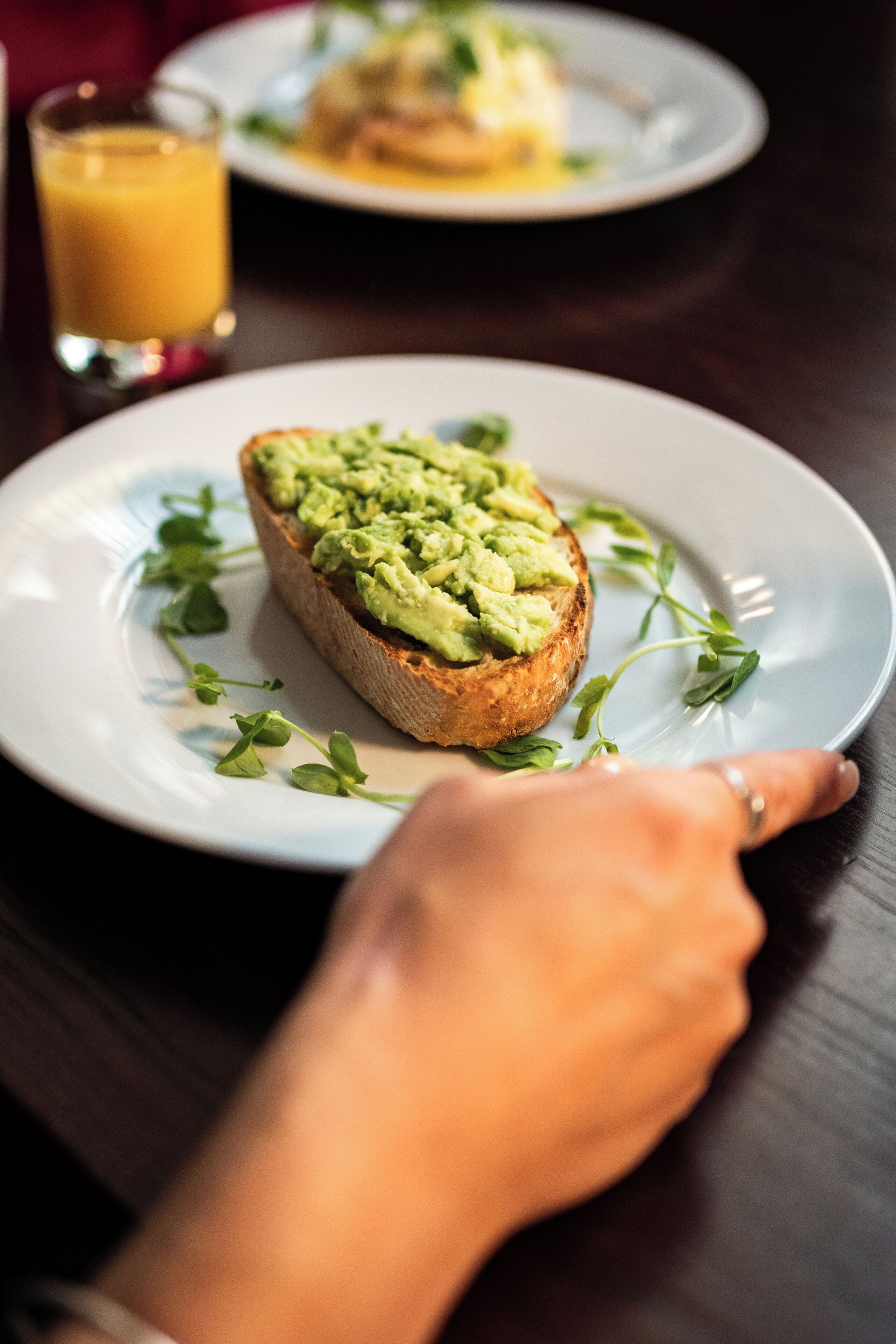 Un plato con tostada de aguacate sobre un plato blanco, un vaso de jugo de naranja y una mano señalando la tostada.