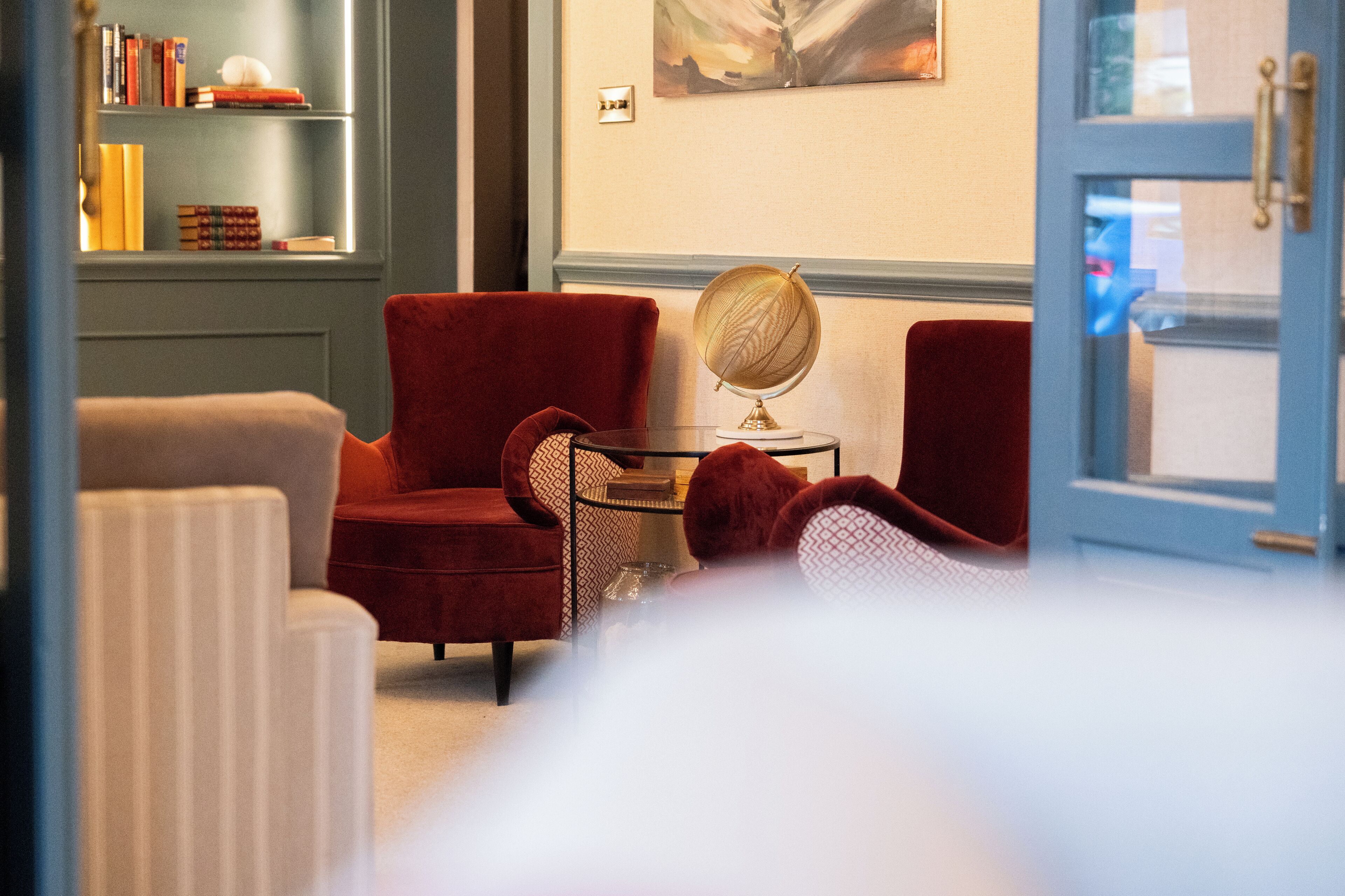 Ein Hotelzimmer mit einem roten Sessel, einem kleinen runden Tisch mit Globus, einem Bücherregal mit Büchern und einem Gemälde an der Wand.