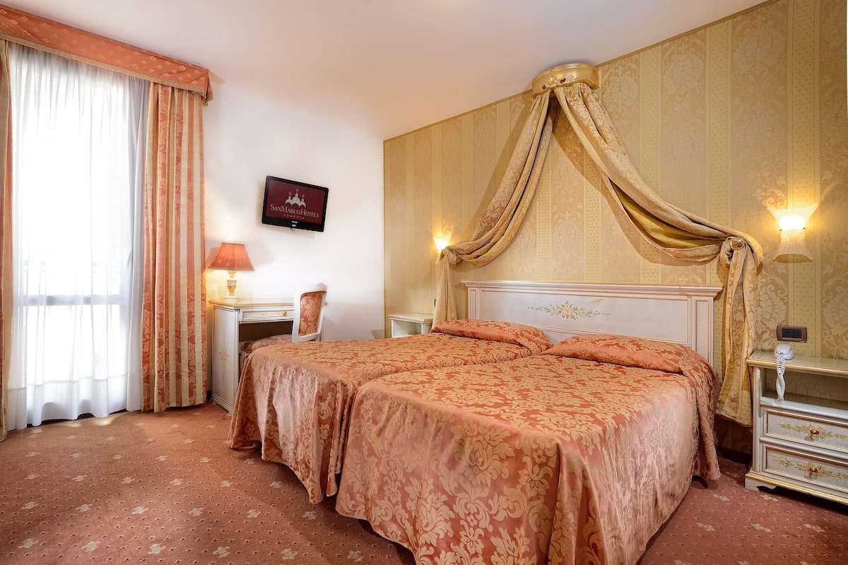 A double bed with a canopy, a bedside table, a lamp, and a chair in a room with patterned wallpaper and a large window with curtains.