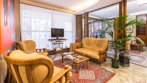 A living room with a sofa, armchairs, a coffee table, a television, and a large window with sheer curtains. - Hotel Ambassador Tre Rose (Venice)