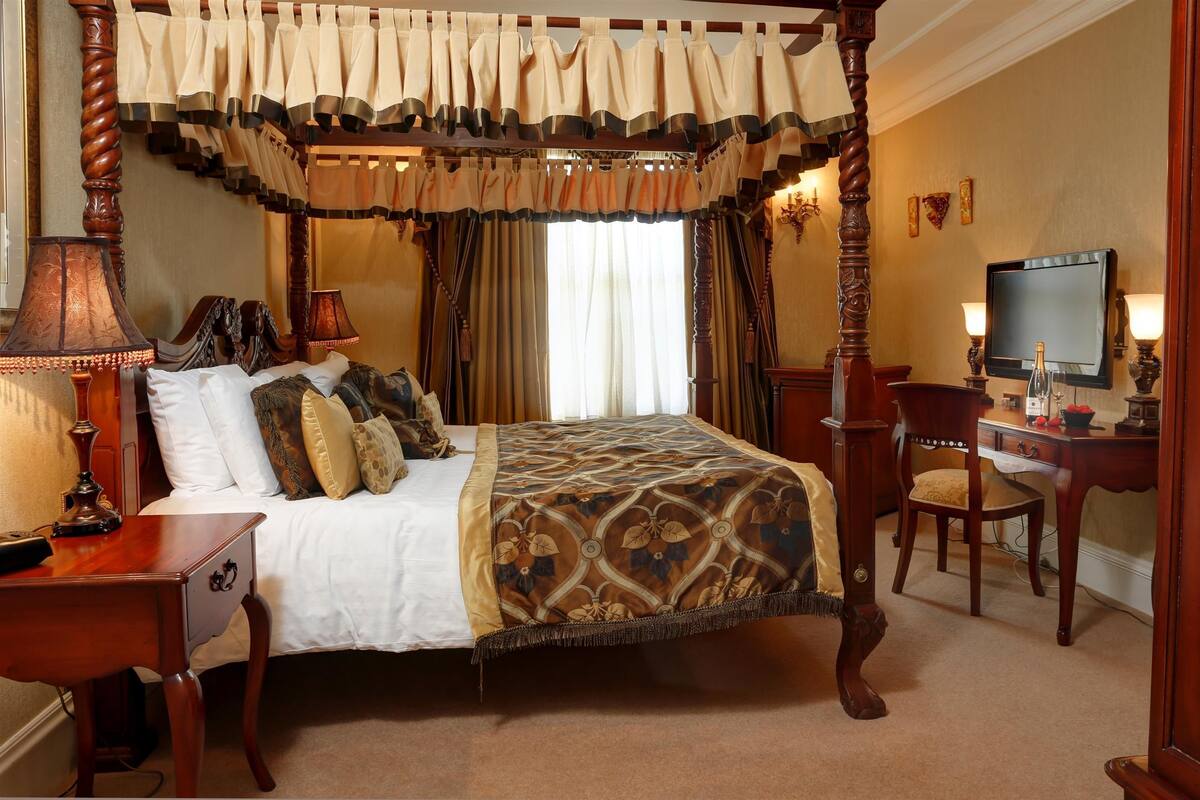 A four-poster canopy bed with a patterned bedspread, a wooden nightstand with a lamp, a desk with a chair, and a television on the wall.