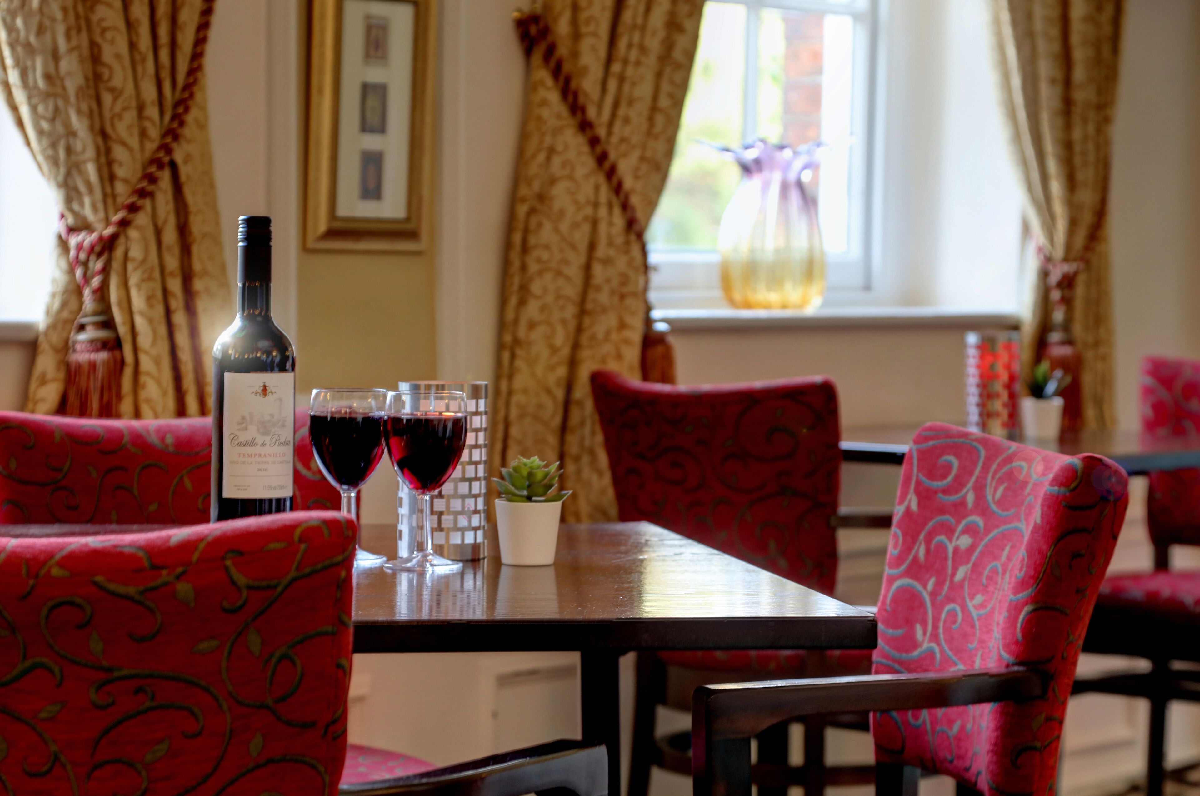 A dining table with a bottle of wine and two glasses of red wine, surrounded by patterned chairs.
