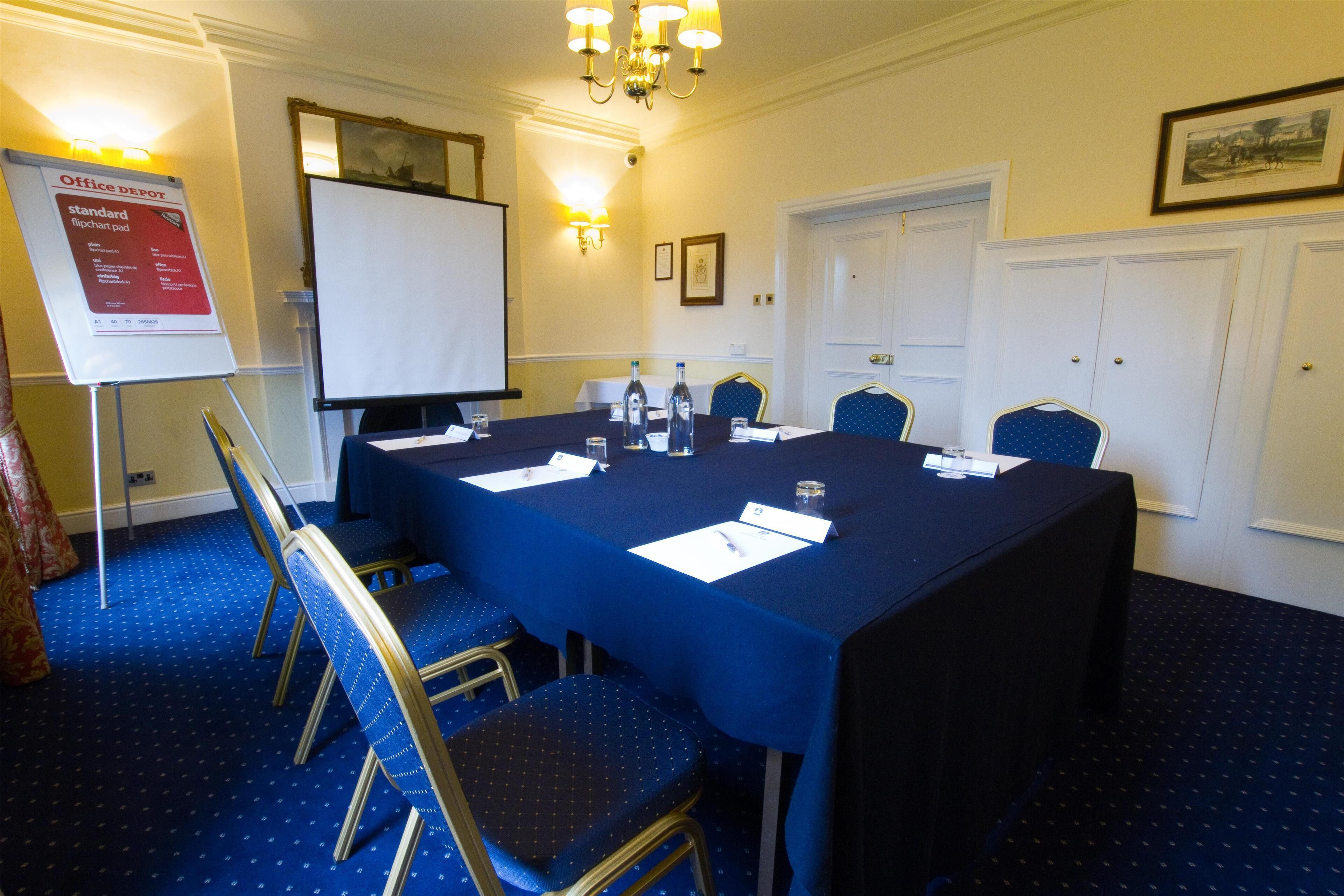 A conference room with a long table, chairs, a projector screen, and a sign about office supplies.