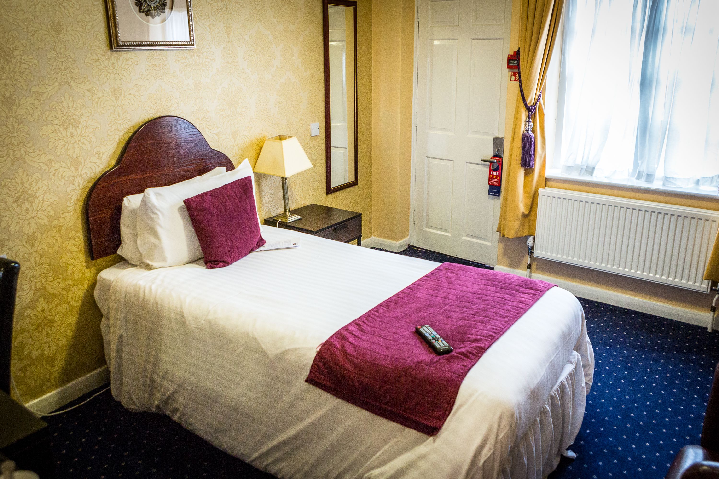 A hotel room with a neatly made bed, a bedside table with a lamp, a mirror, a door, and a window with curtains.