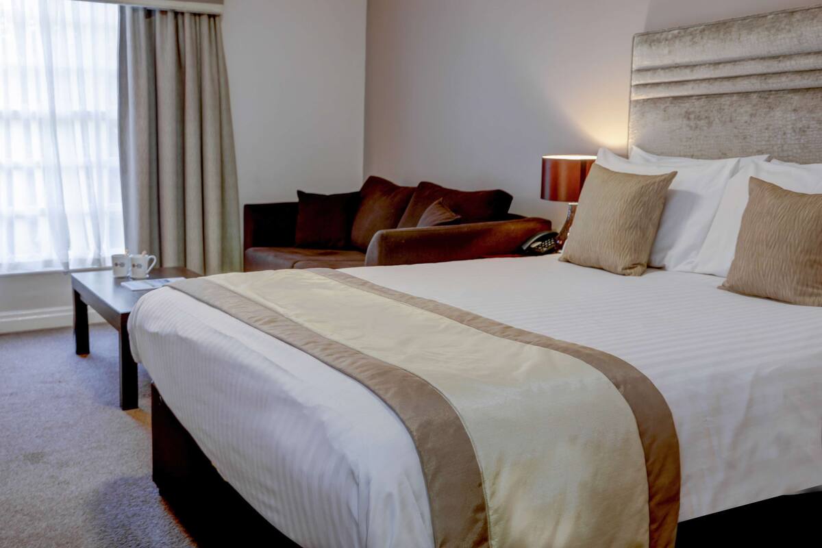 A hotel room with a large bed, a bedside table, a lamp, a sofa, and a window with curtains.