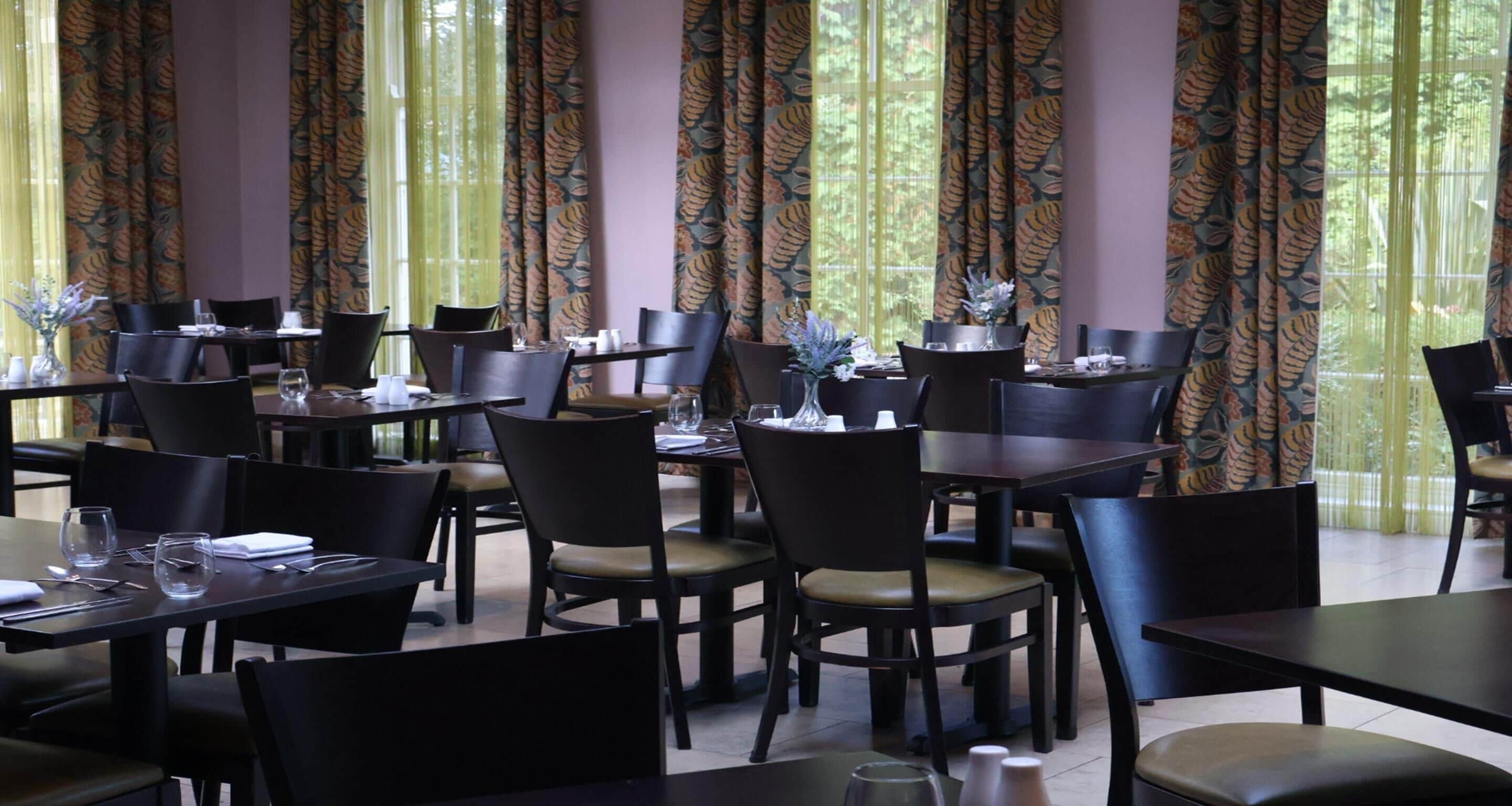 Un restaurant doté de nombreuses tables et chaises, de grandes fenêtres avec des rideaux et offrant une vue sur la verdure environnante.