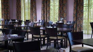 A restaurant with multiple tables and chairs, large windows with curtains, and a view of greenery outside.