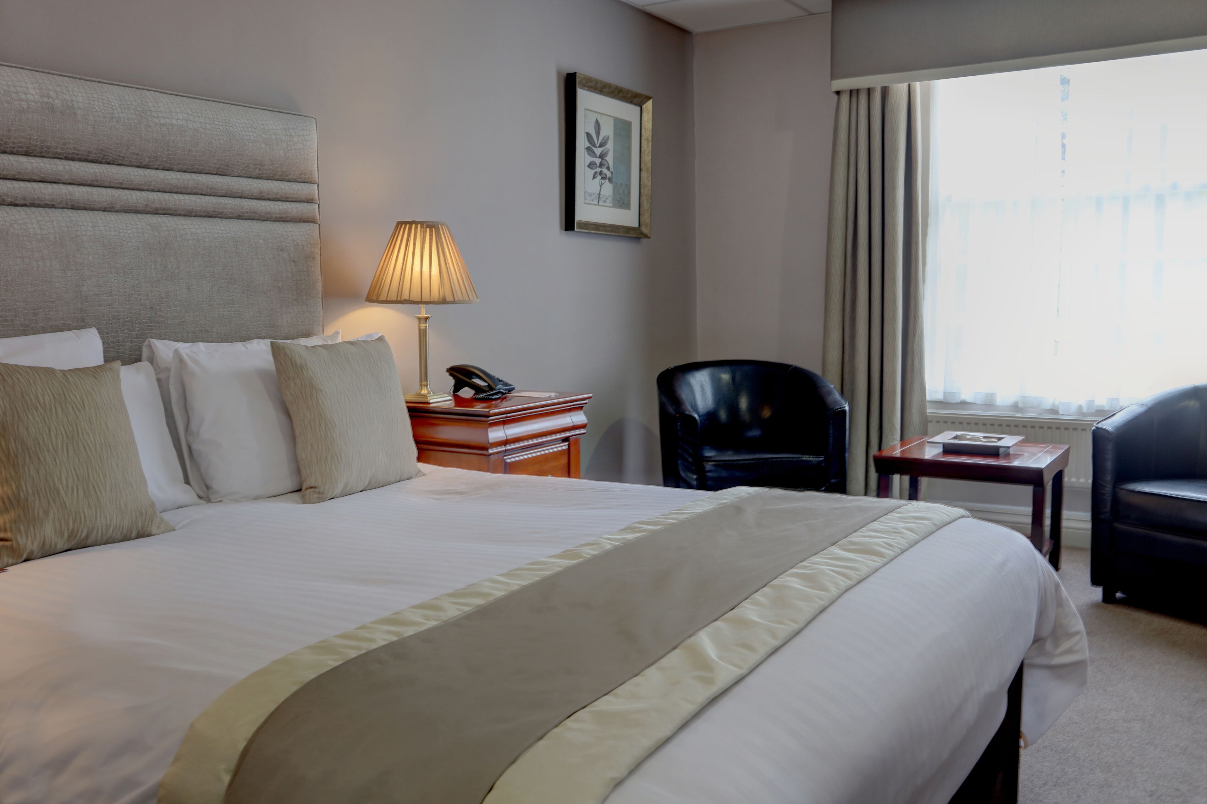 A hotel room with a large bed, a bedside table with a lamp, a chair, and a window with curtains.