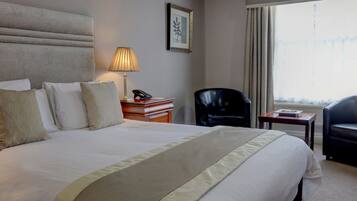Ein Hotelzimmer mit einem großen Bett, einem Nachttisch mit Lampe, einem Sessel und einem Fenster mit Vorhängen.