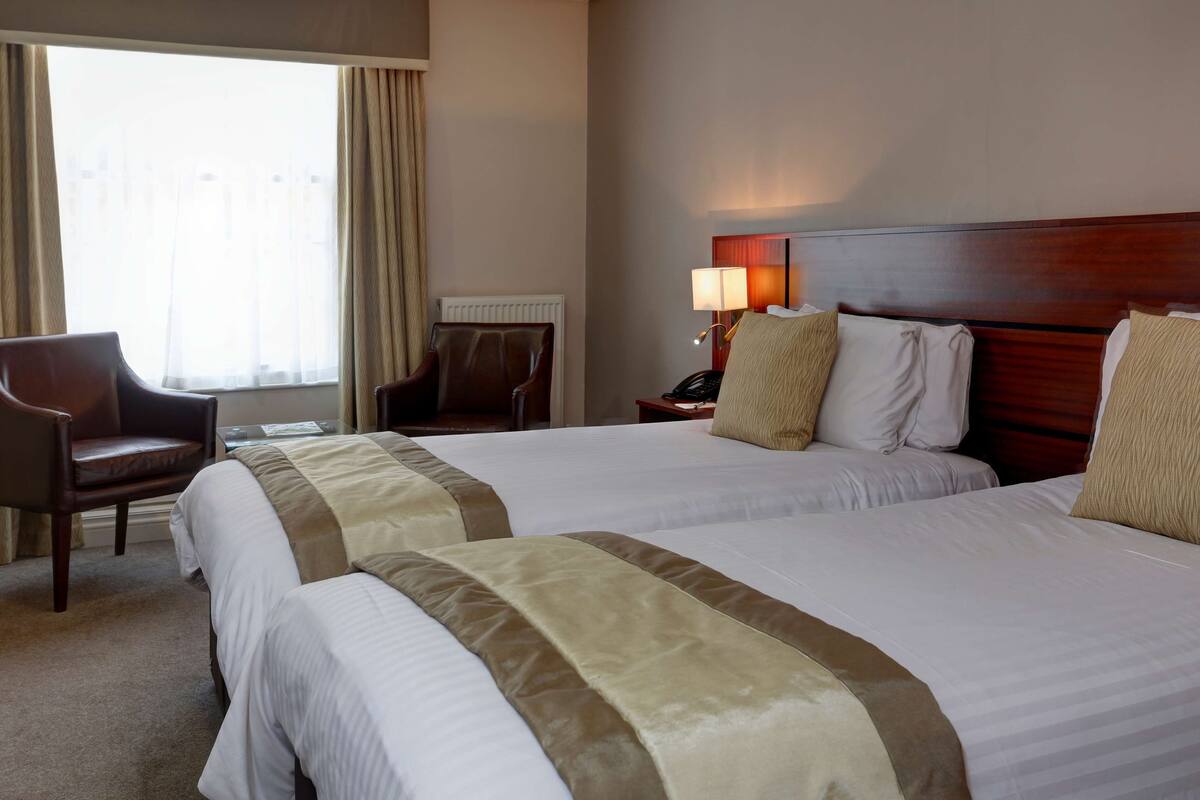 A hotel room with two beds, a chair, a lamp, and a window with curtains.