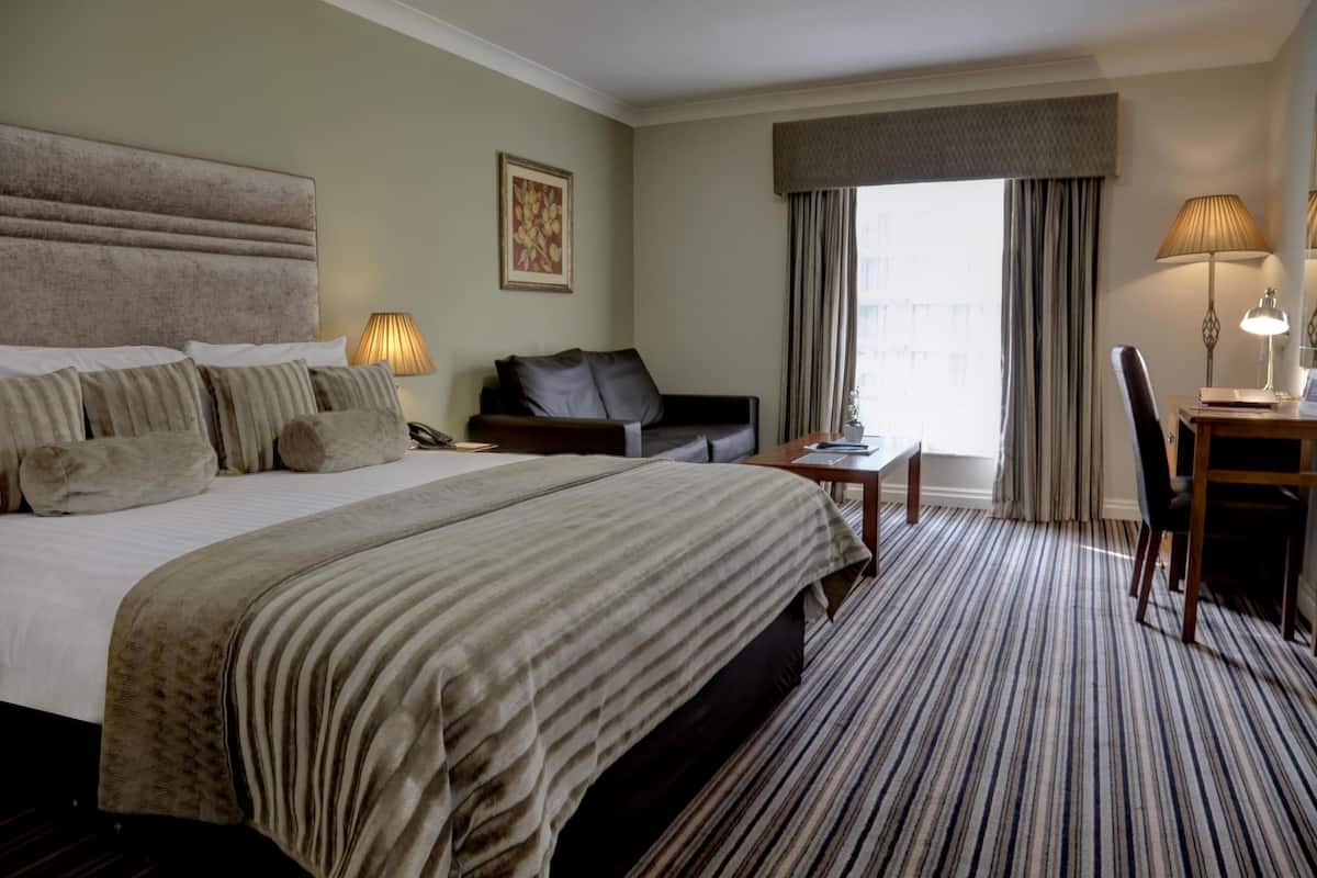 A hotel room with a large bed, a sofa, a desk with a chair, and a window with curtains.
