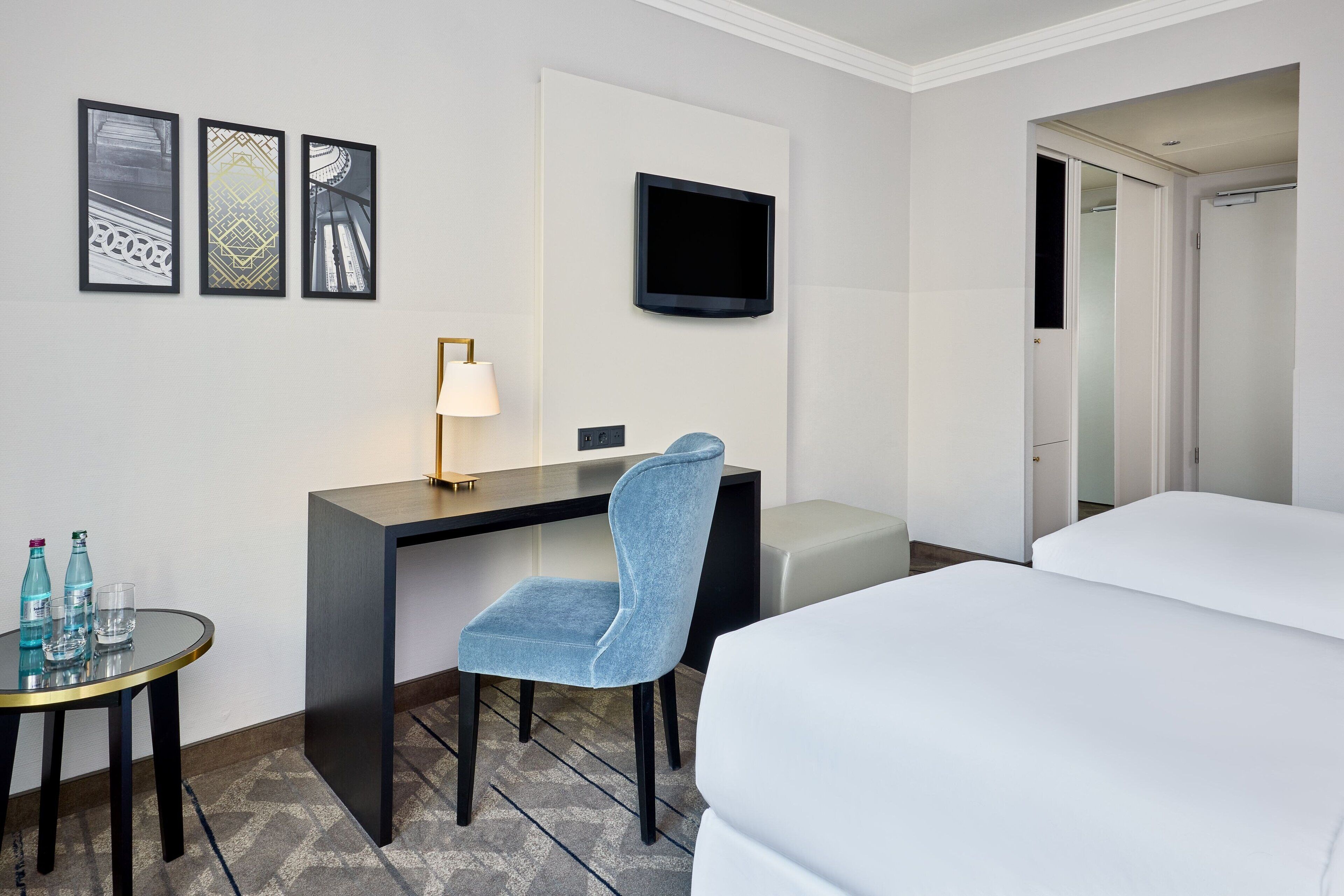 A hotel room with a bed, a desk with a chair, a TV, and a small table with water bottles.