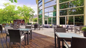 An outdoor patio with wicker furniture, large windows, and a view of trees and a building in the background. - Sheraton Offenbach Hotel (Offenbach am Main)