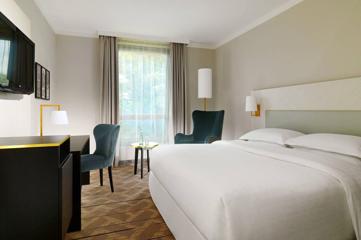 Ett hotellrum med en säng, ett skrivbord, en stol, en tv och ett fönster med gardiner.