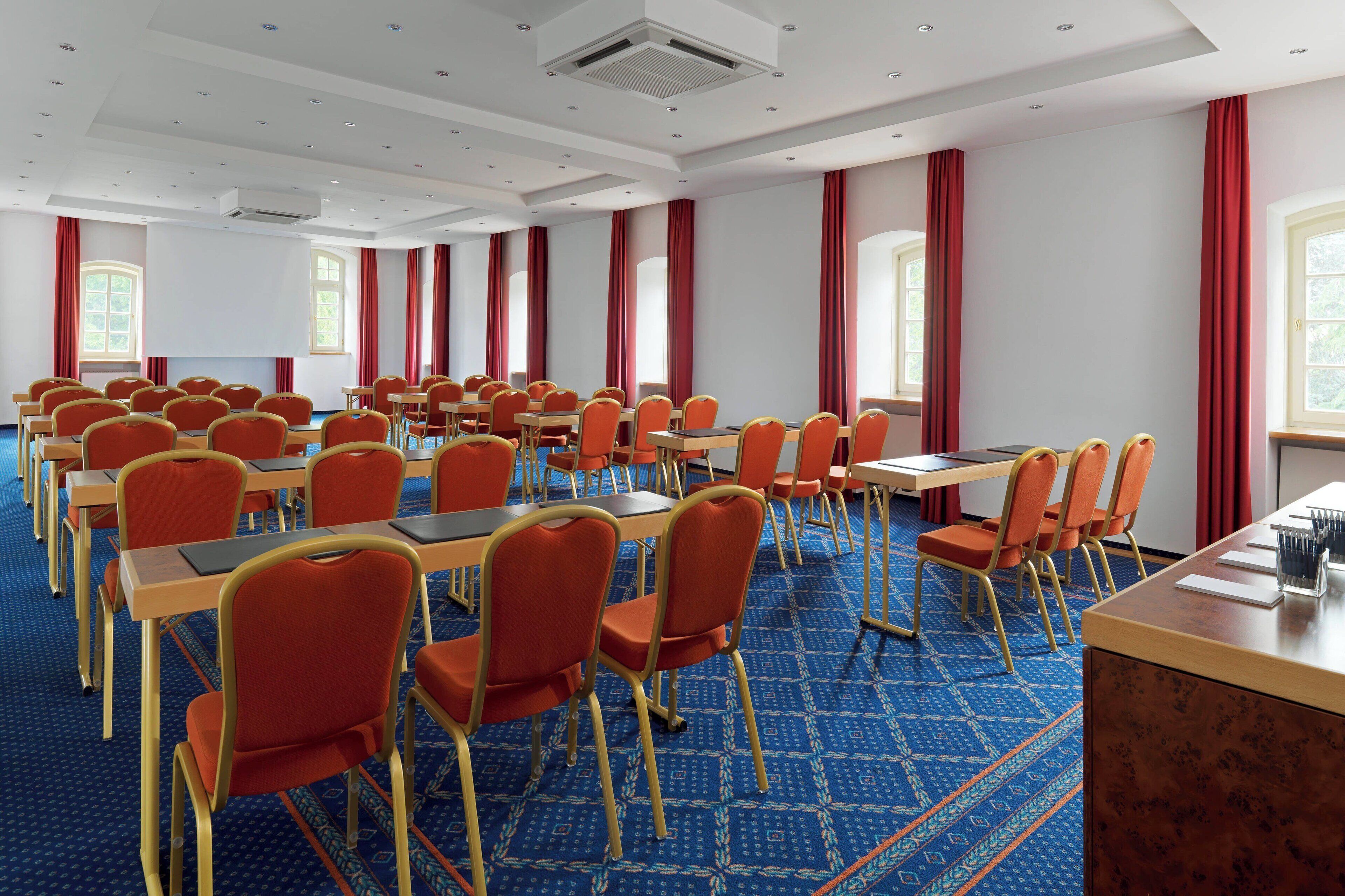 Un salón de conferencias con filas de mesas y sillas, ventanas amplias con cortinas rojas y una alfombra estampada.