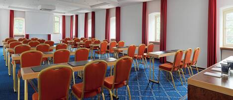 Un salón de conferencias con filas de mesas y sillas, ventanas amplias con cortinas rojas y una alfombra estampada.
