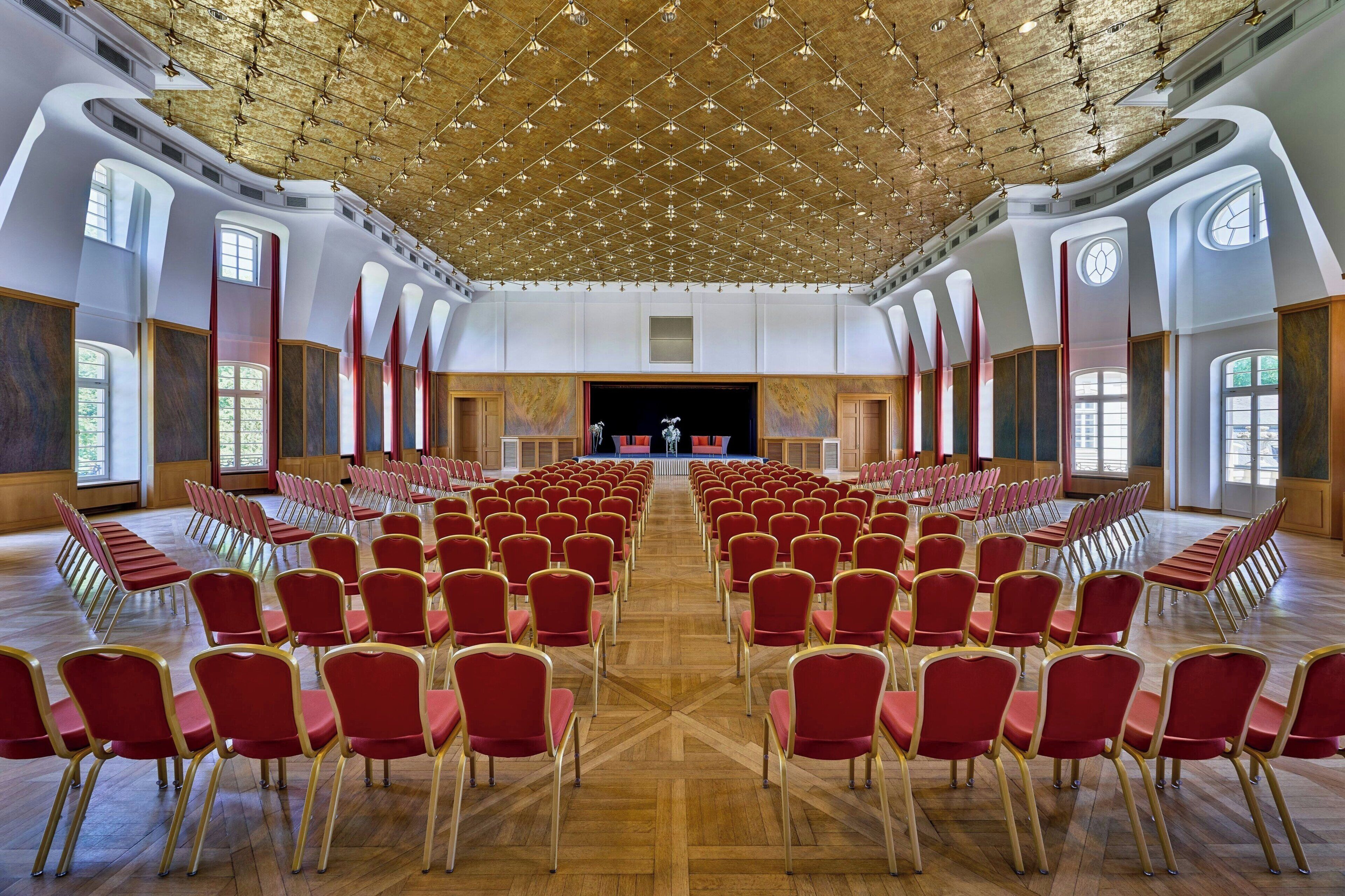 A large hall with rows of red chairs and a stage at the front.