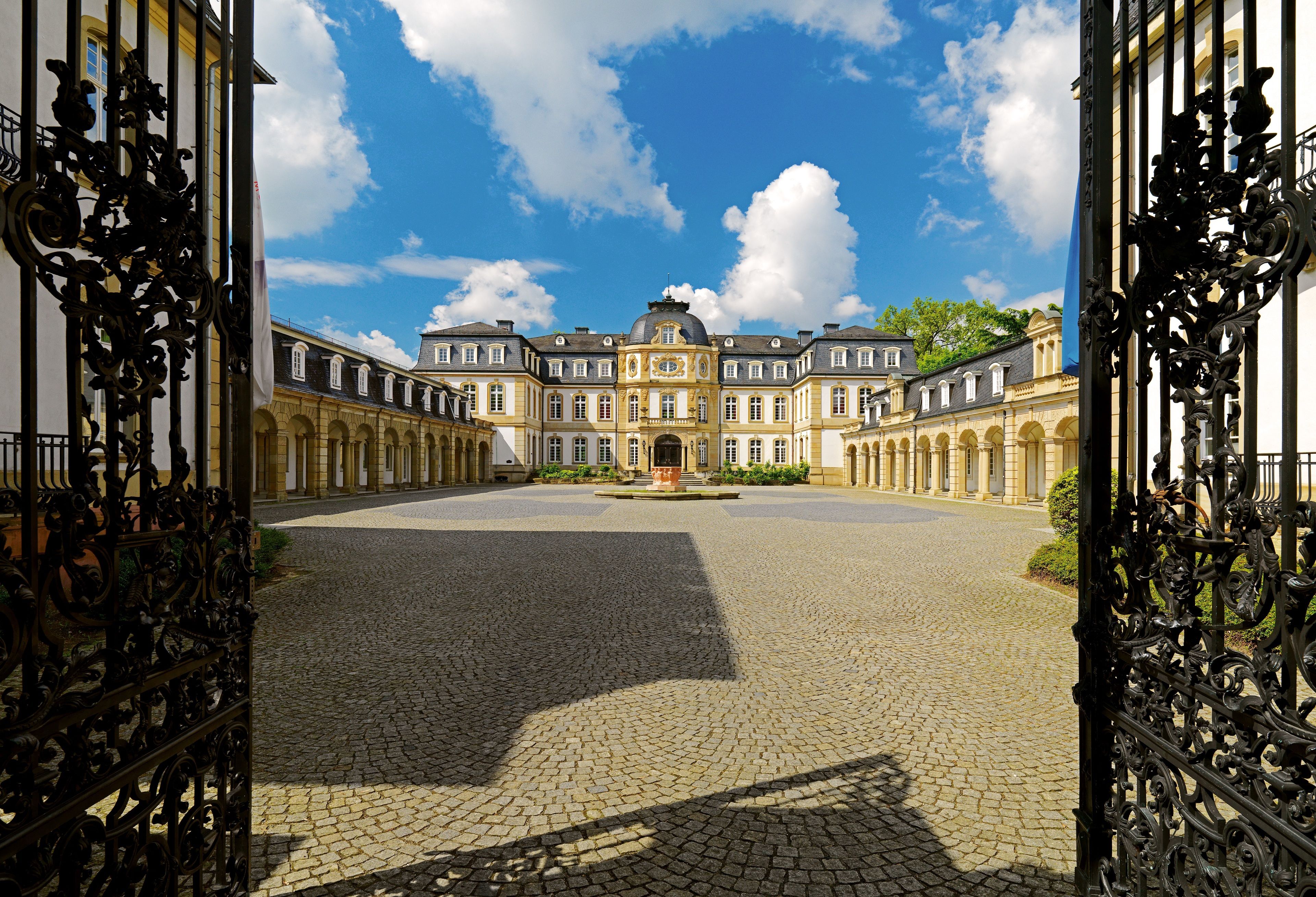 A courtyard with a large building in the background, featuring a central fountain and symmetrical architecture.
