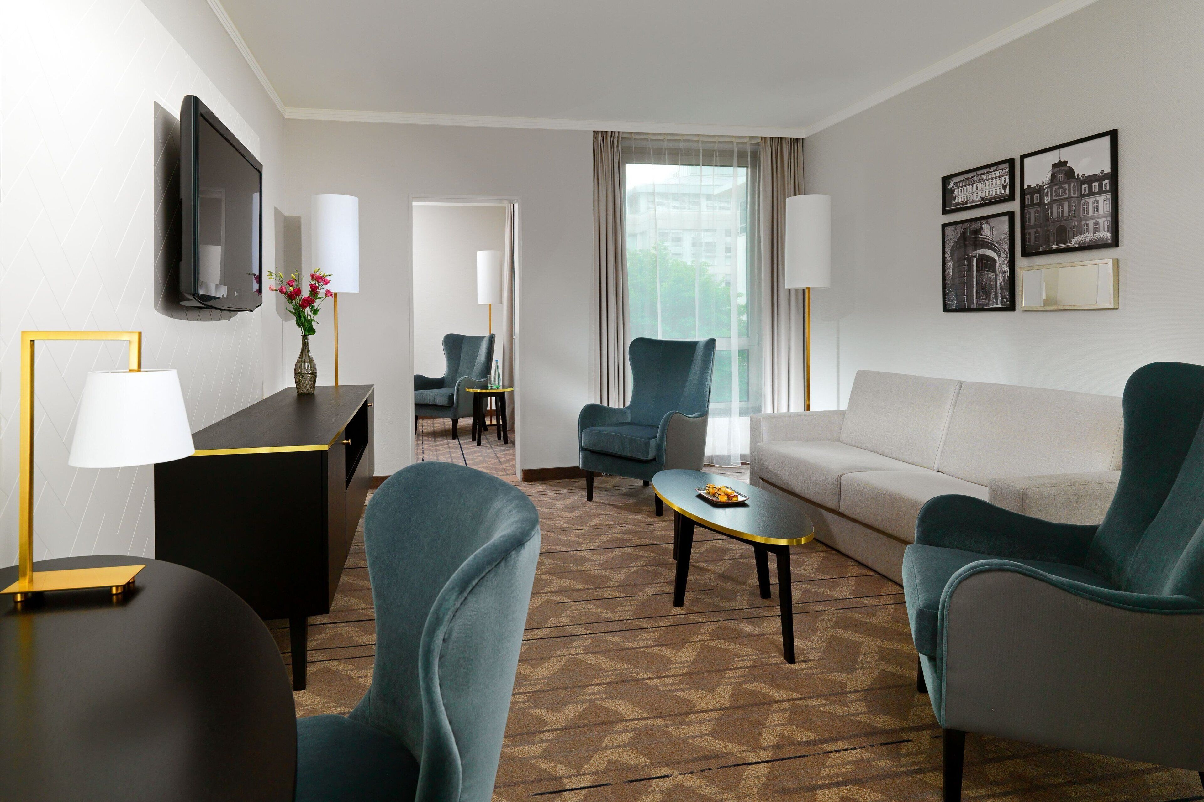 Una habitación de hotel moderna con sofá, dos sillones, una mesa de centro, un televisor montado en la pared y una zona de comedor con sillas y mesa.