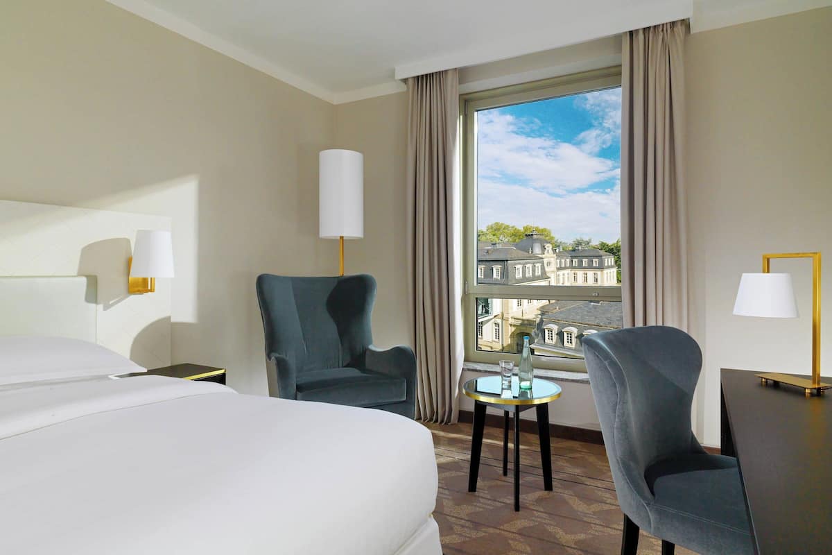 Ett hotellrum med en stor säng, två fåtöljer, ett litet bord och utsikt över byggnader genom ett fönster.
