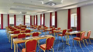 Un salón de conferencias con filas de mesas y sillas, un ventanal con cortinas rojas y un logotipo en la pared.