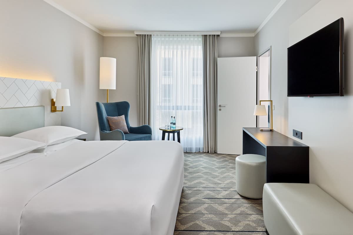 Ett hotellrum med en stor säng, ett skrivbord, en stol, en TV och ett fönster med gardiner.
