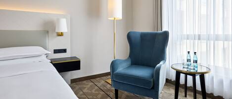 Une chambre d’hôtel avec un lit, un fauteuil bleu, une petite table avec des bouteilles d’eau, une lampe sur pied et des rideaux légers.