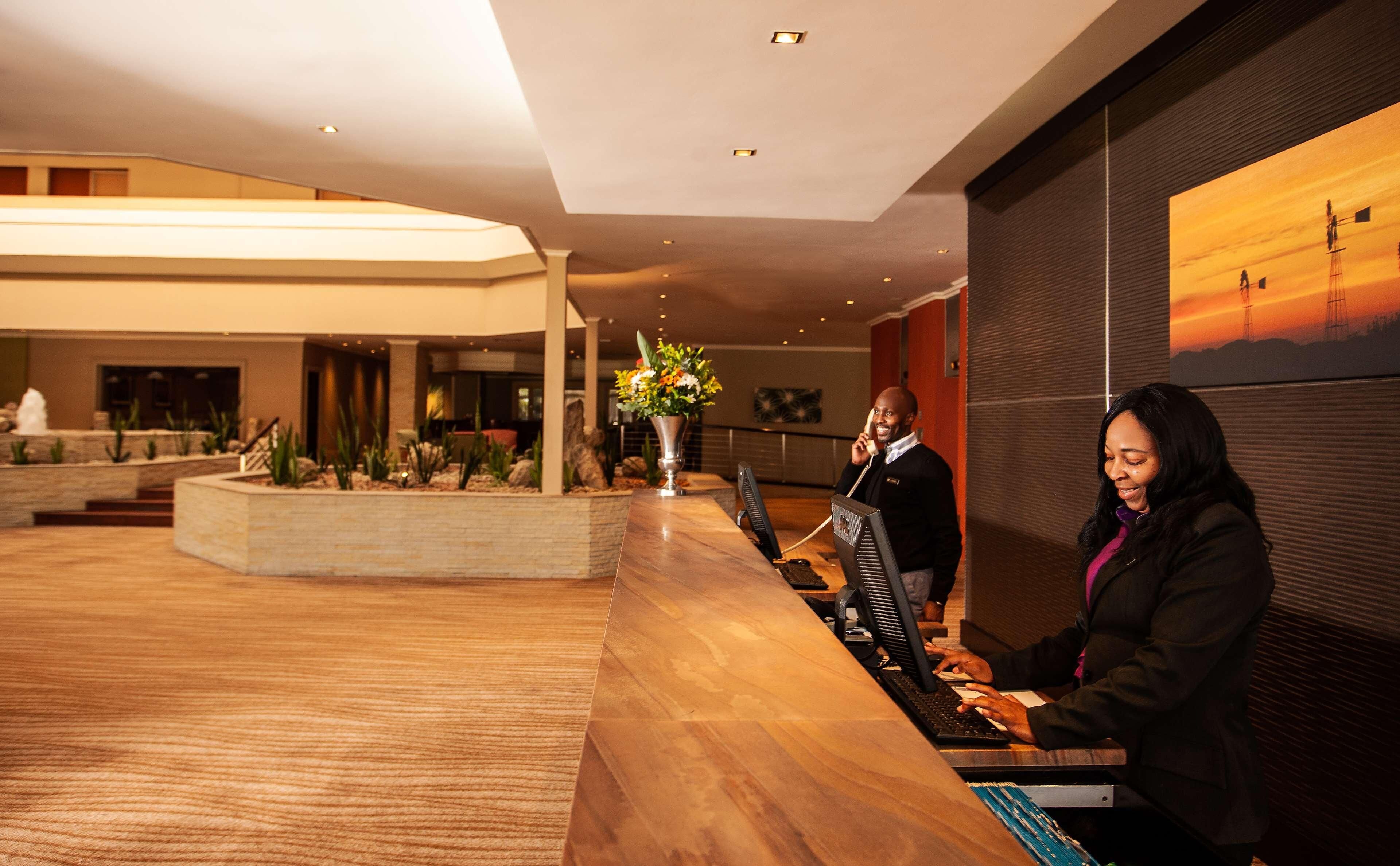 A reception desk with a computer, a vase with flowers, and a painting of a sunset on the wall.