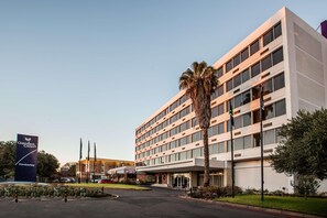Ein mehrstöckiges Hotel mit modernem Design, das einen großzügigen Eingangsbereich, einen Parkplatz und eine markante Beschilderung bietet.