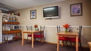 A dining area with two wooden tables and red chairs, a television mounted on the wall, and a shelf with kitchen items.
