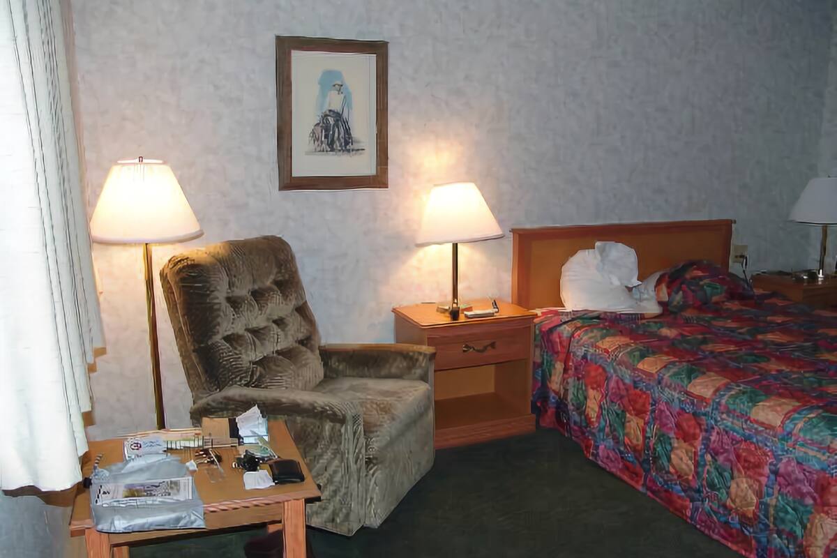 A hotel room with a large armchair, a bedside table with a lamp, a bed with a colorful blanket, and a framed picture on the wall.