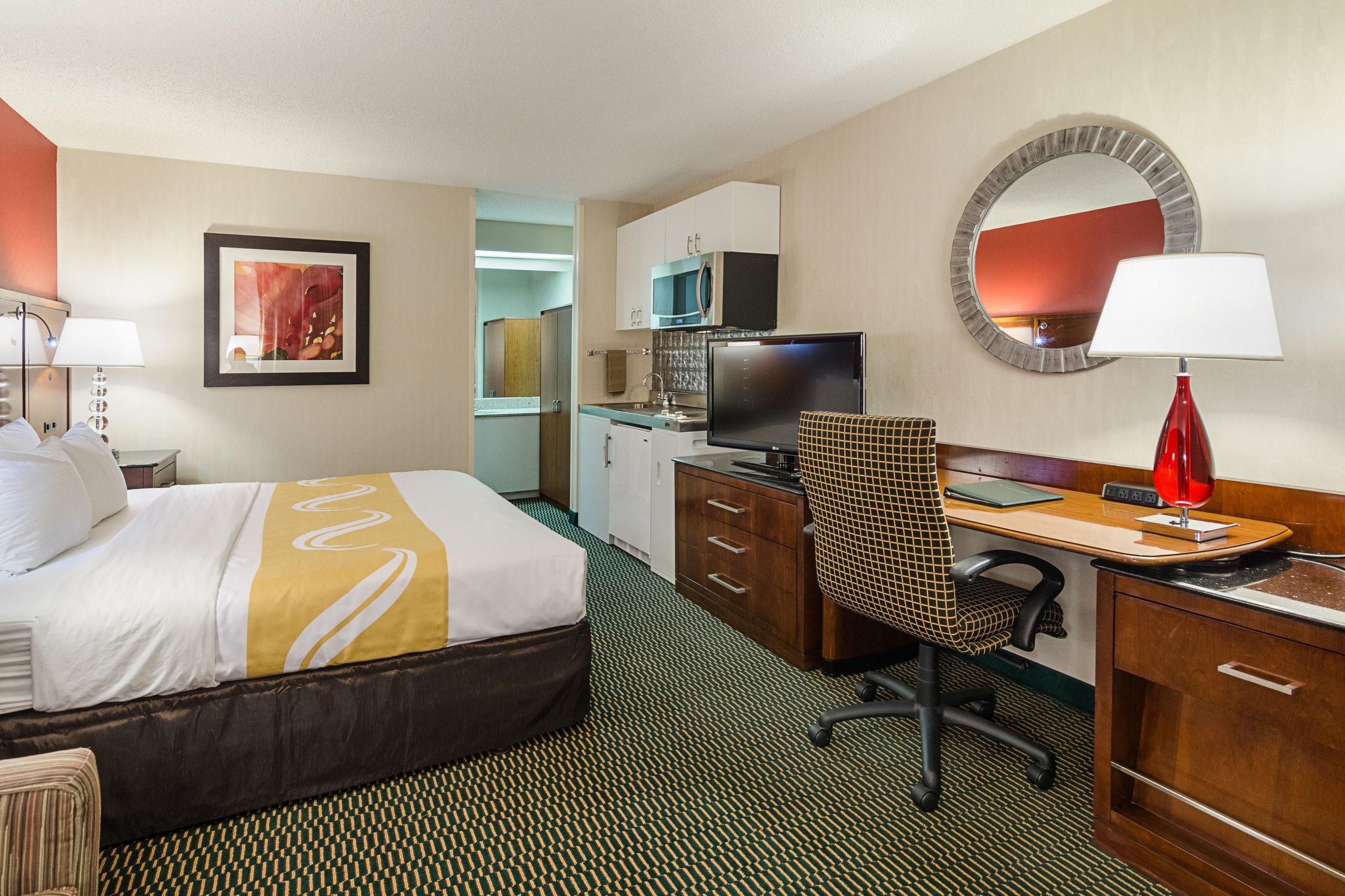 Habitación de hotel con cama, escritorio, silla, televisor, microondas y espejo.