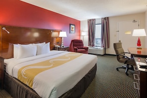 Una habitación de hotel con una cama grande, un escritorio con silla, un sillón rojo y una ventana con cortinas.
