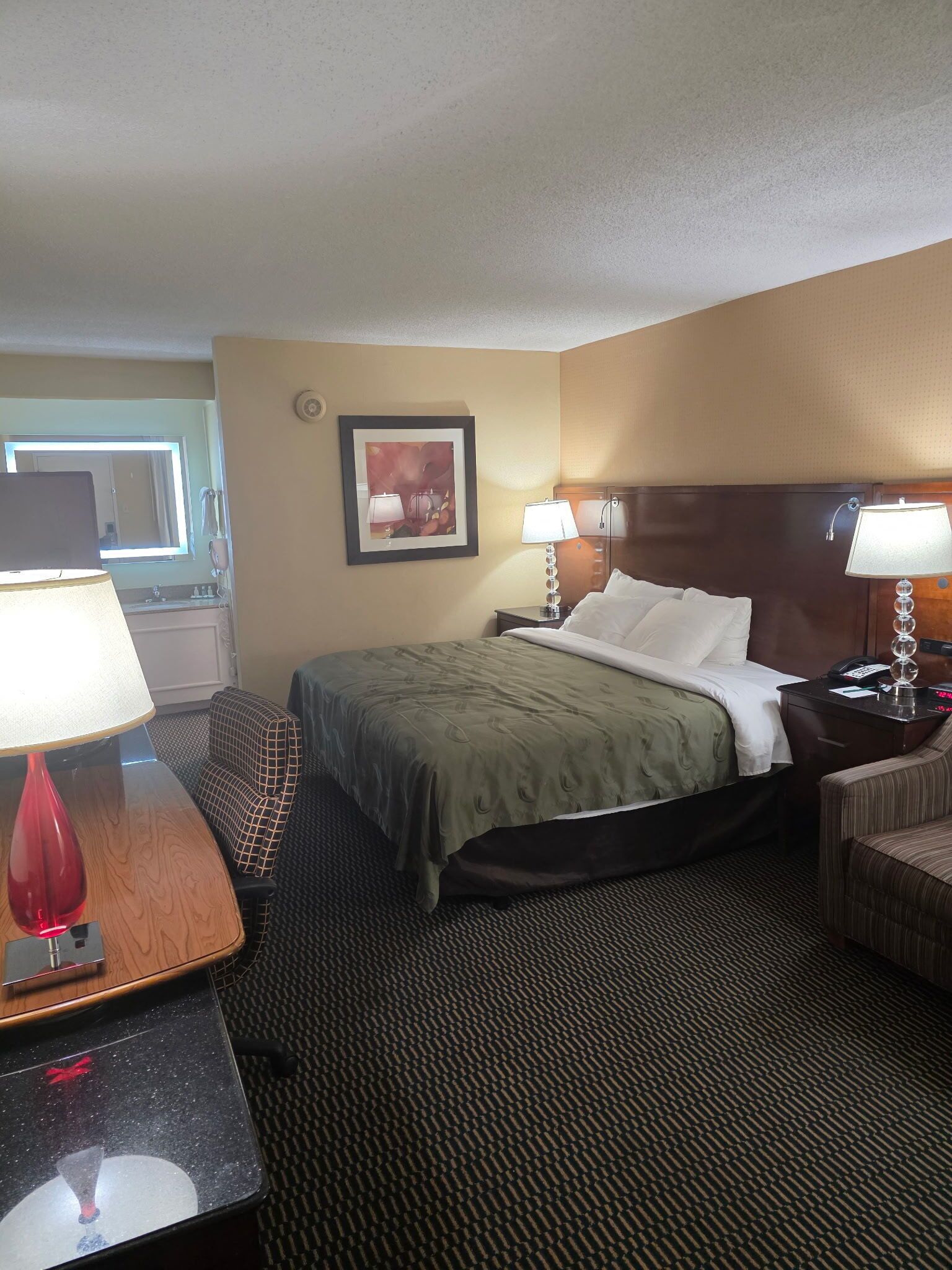 A hotel room with a bed, two bedside tables, a desk, a chair, a lamp, a framed picture, and a window.