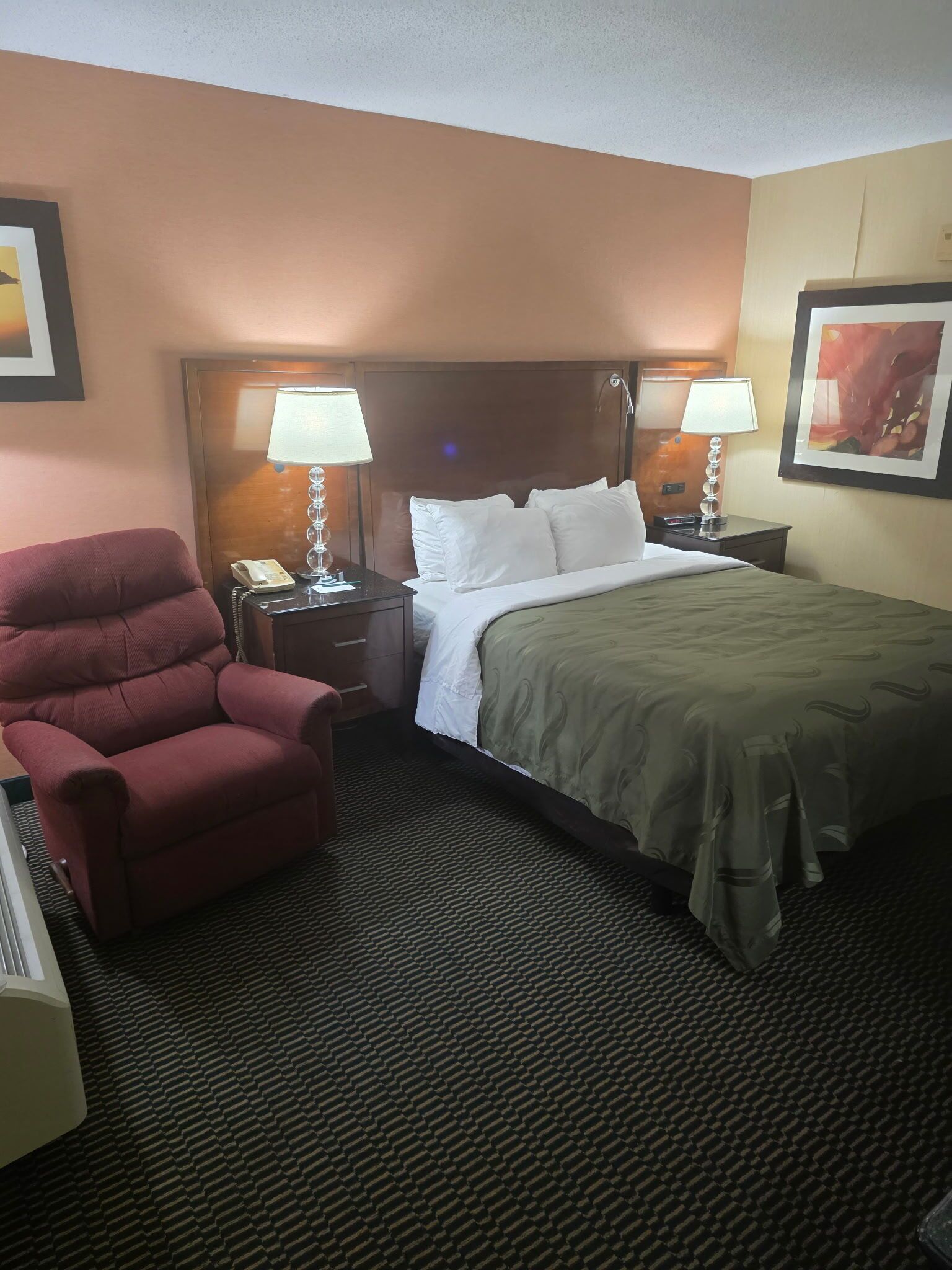 Une chambre d’hôtel avec un lit, deux lampes de chevet, un fauteuil rouge, un téléphone et un tableau encadré au mur.
