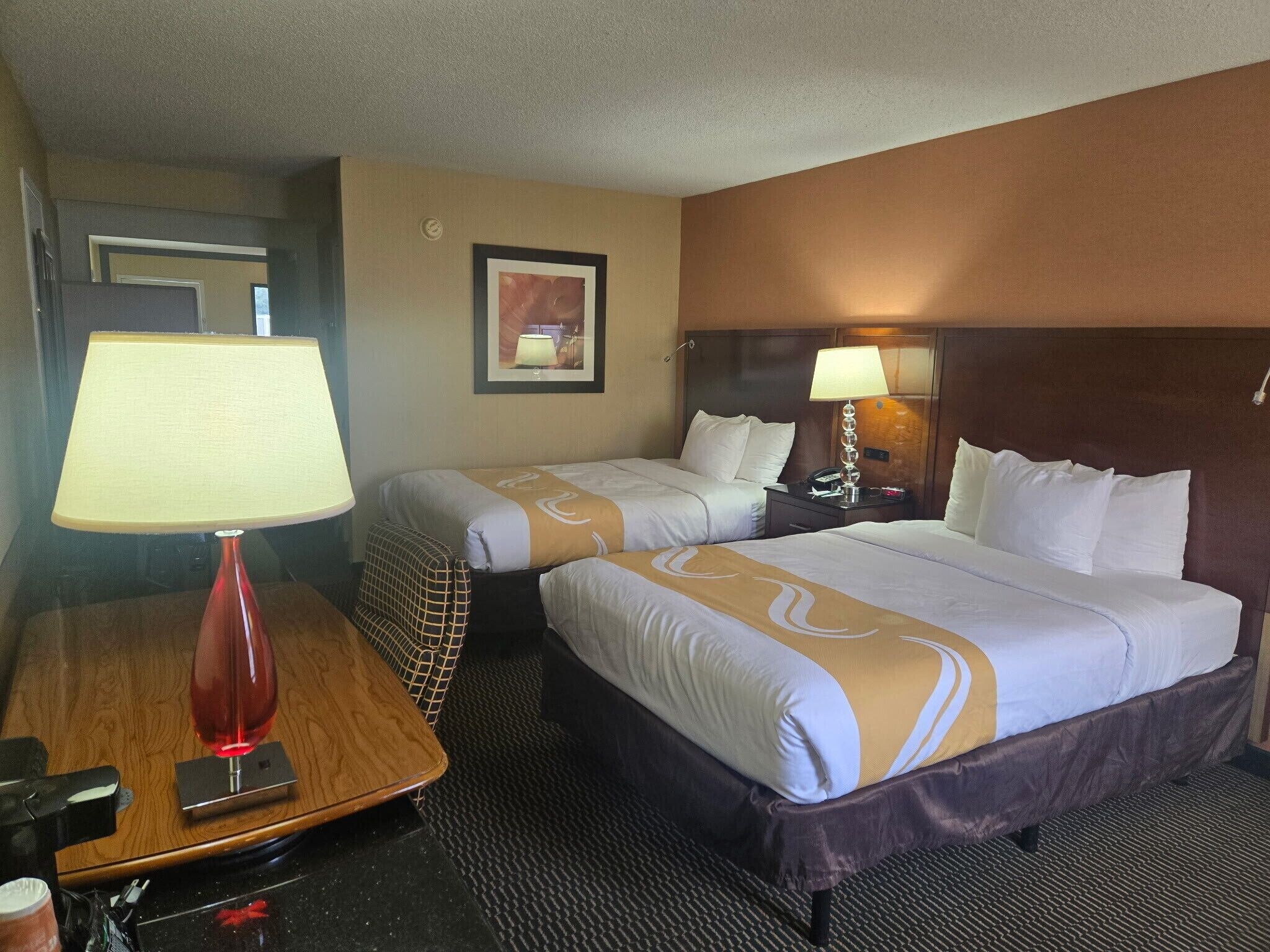 Une chambre d’hôtel avec deux lits, un bureau, une lampe rouge et un tableau encadré au mur.
