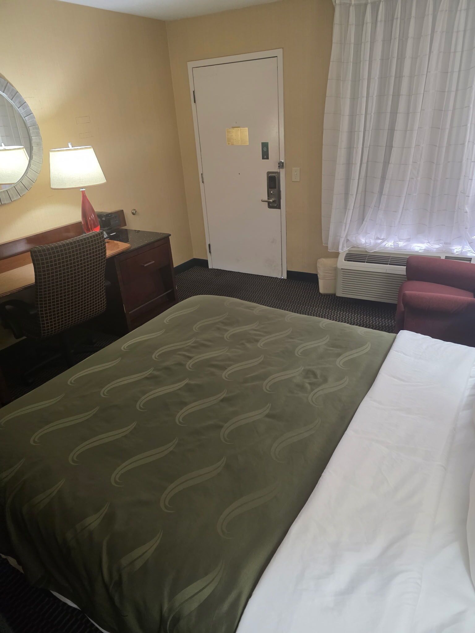 A hotel room with a bed, a desk with a lamp, a chair, a door, and a window with curtains.