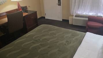 A hotel room with a bed, a desk with a lamp, a chair, a door, and a window with curtains.