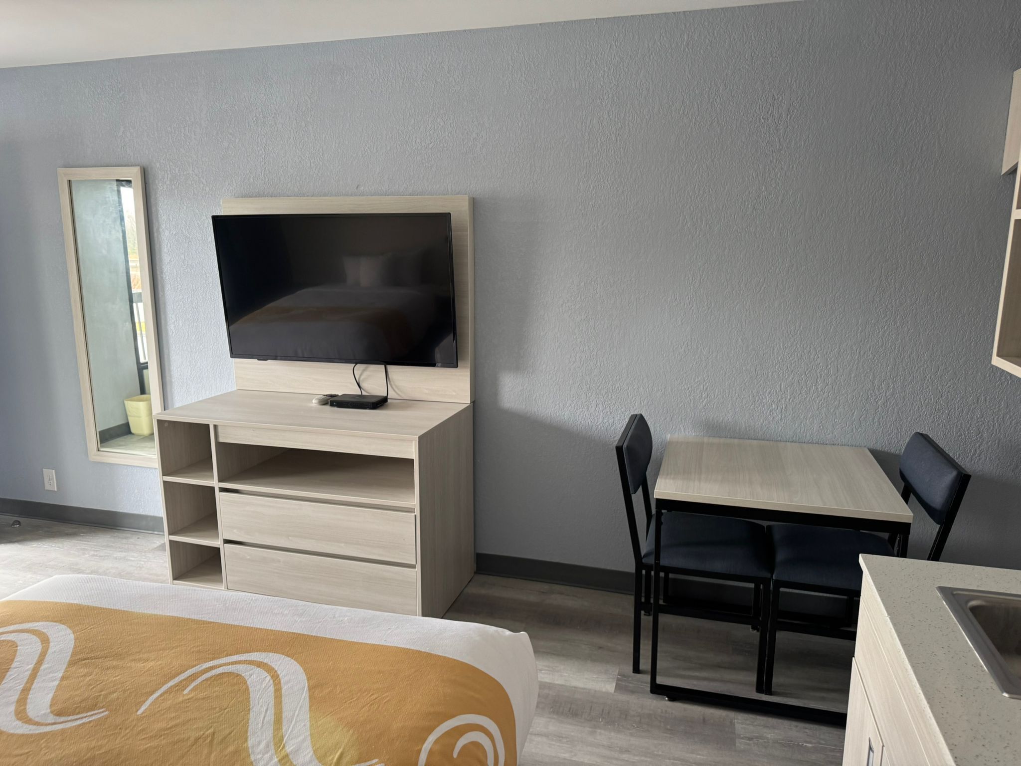 Una habitación de hotel moderna con una televisión de pantalla plana sobre un mueble de madera, una mesa de comedor con sillas, un espejo grande y una cama con cubrecama estampado.