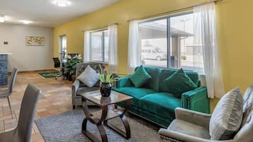 Una sala de estar con un sofá verde, una mesa de centro y una silla. Hay una ventana con cortinas, una puerta y una planta sobre la mesa.