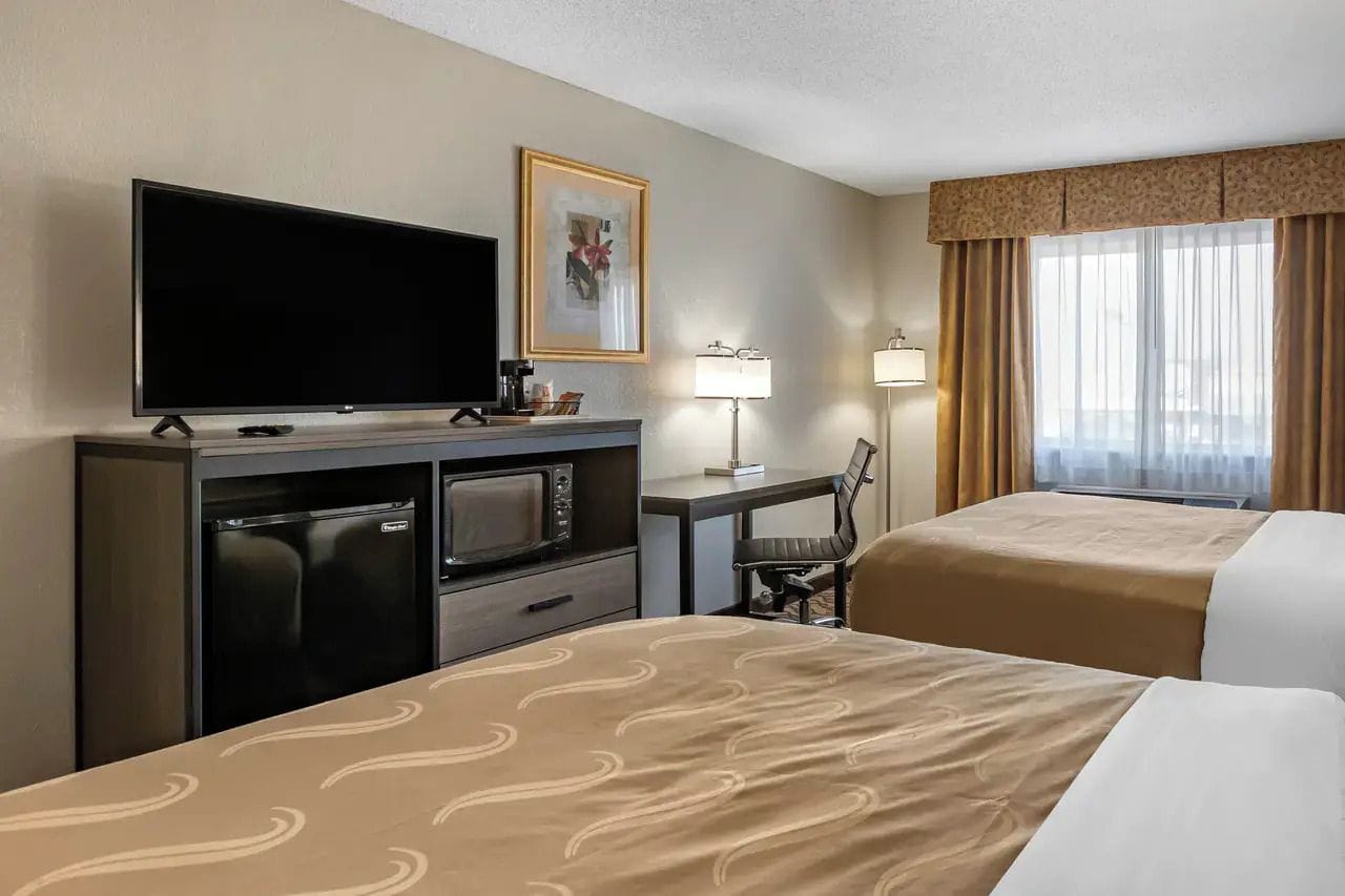 A hotel room with two beds, a television, a desk with a lamp, and a refrigerator.