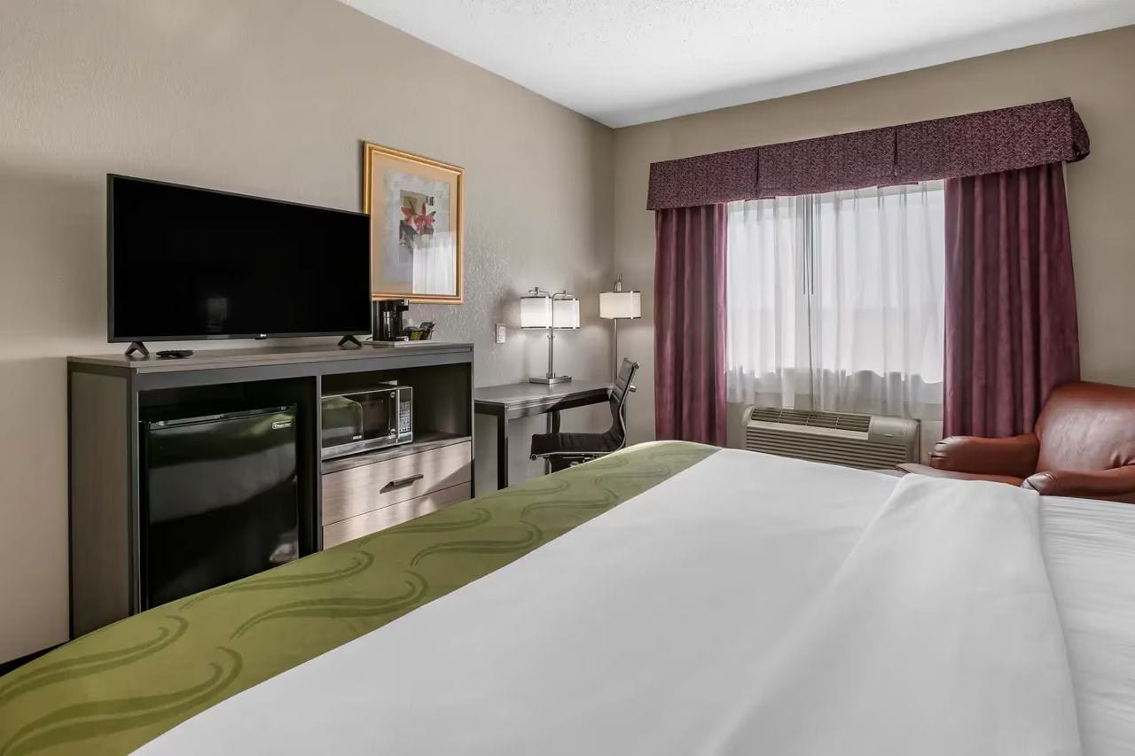 Une chambre d’hôtel équipée d’un lit, d’une télévision, d’un bureau, d’une chaise, d’une lampe et d’une fenêtre avec des rideaux.