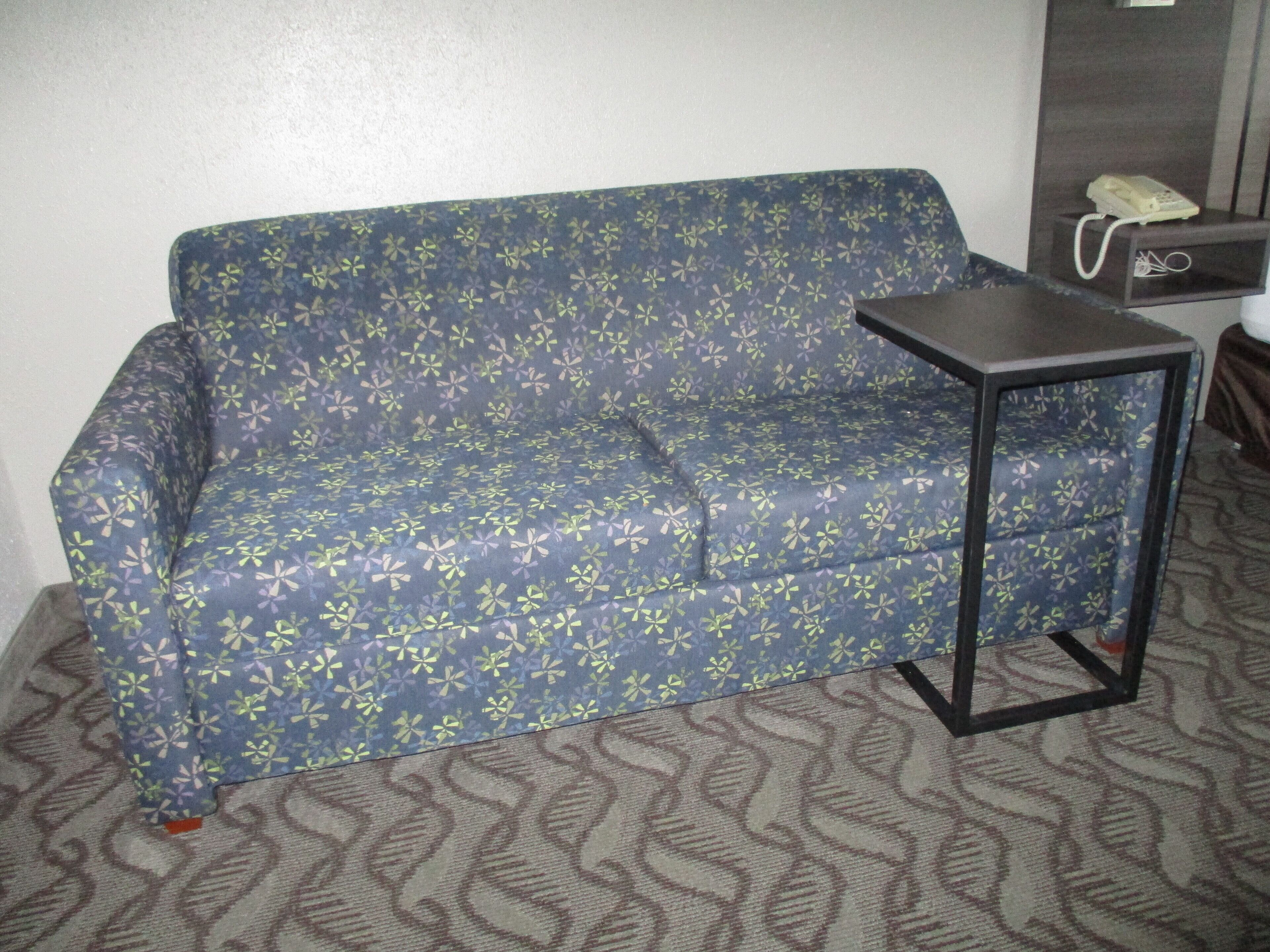 Un sofá azul con estampado floral, una mesita auxiliar negra pequeña y un teléfono sobre un estante de madera.