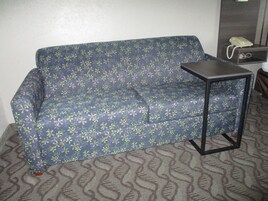 Un sofá azul con estampado floral, una mesita auxiliar negra pequeña y un teléfono sobre un estante de madera.