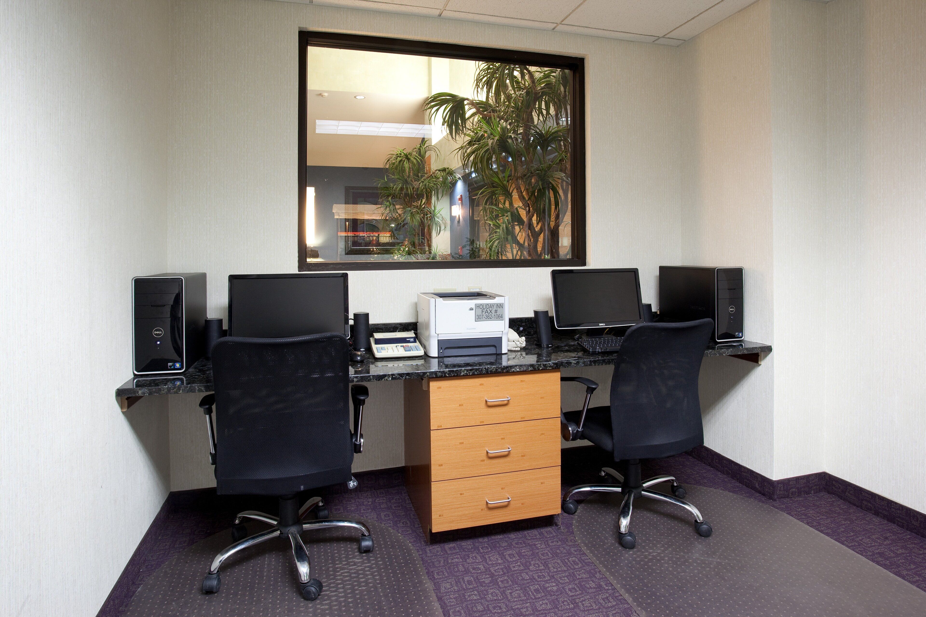 Ein Hotelbüro mit zwei Schreibtischen, zwei Stühlen, einem Drucker und einem Fenster mit Blick auf Palmen.