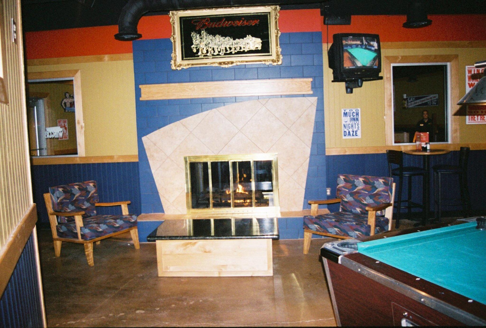 A billiards room with a pool table, two chairs, a fireplace, and a television mounted on the wall.