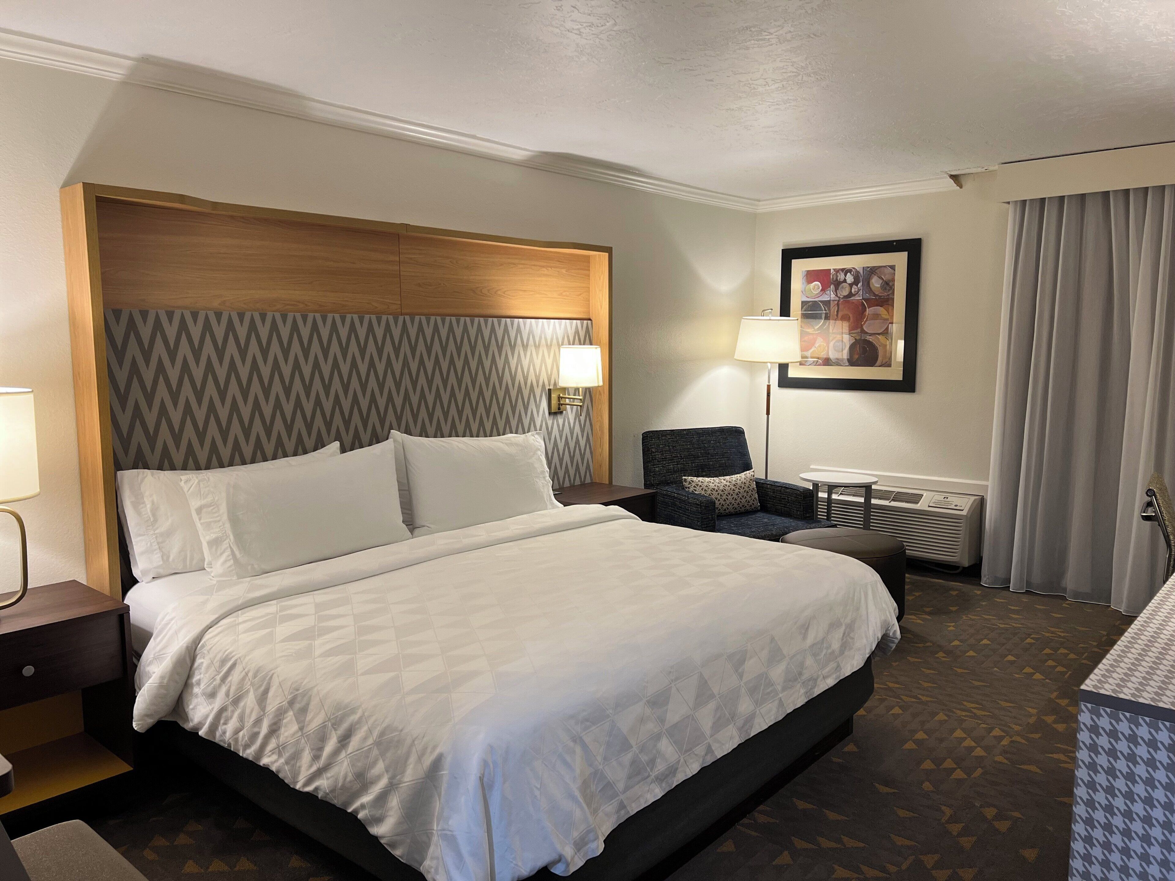 Una habitación de hotel con una cama grande, una silla, una mesita, una lámpara y un radiador.