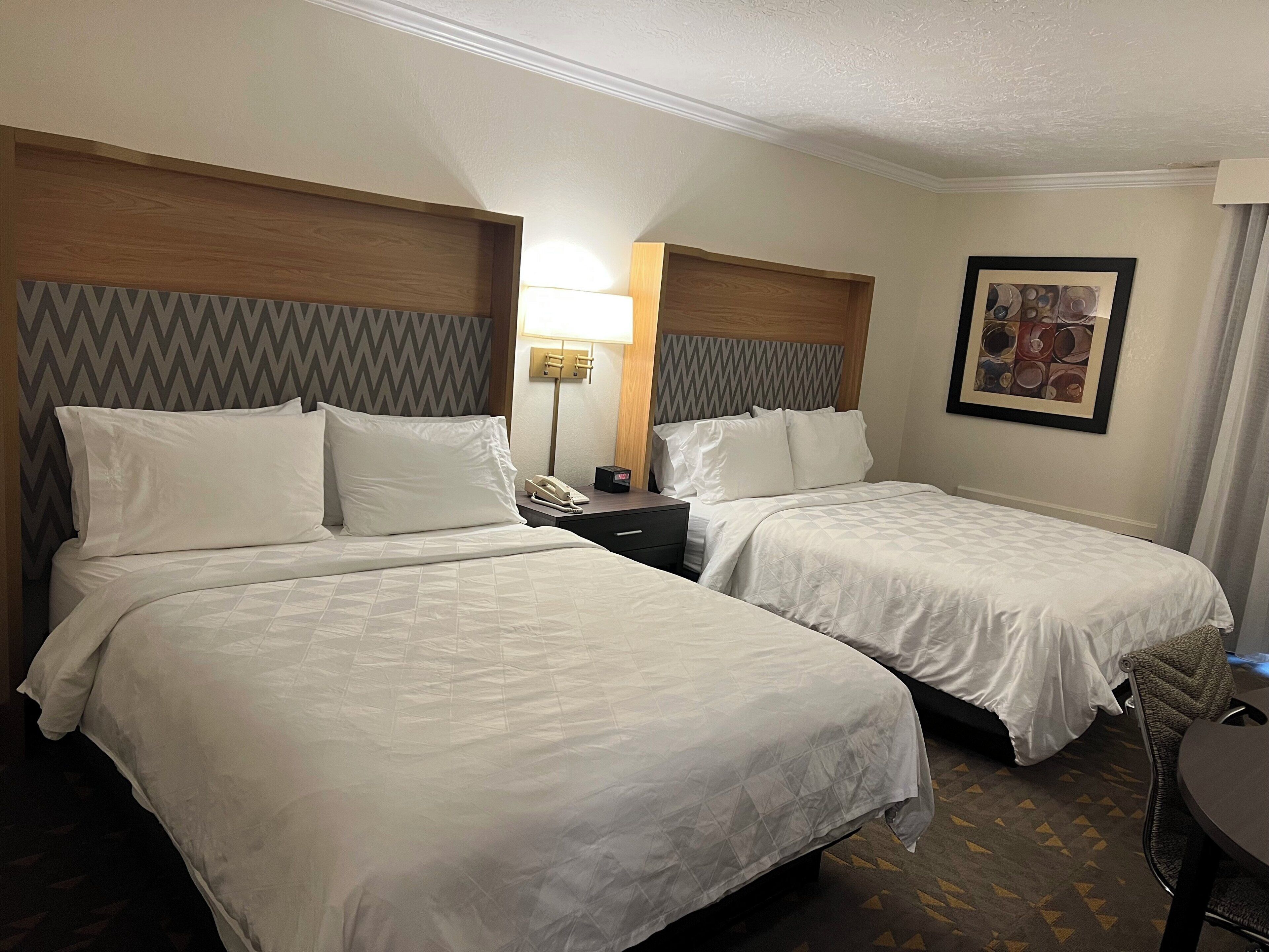 A hotel room with two beds, a bedside table with a lamp, a phone, and a framed picture on the wall.
