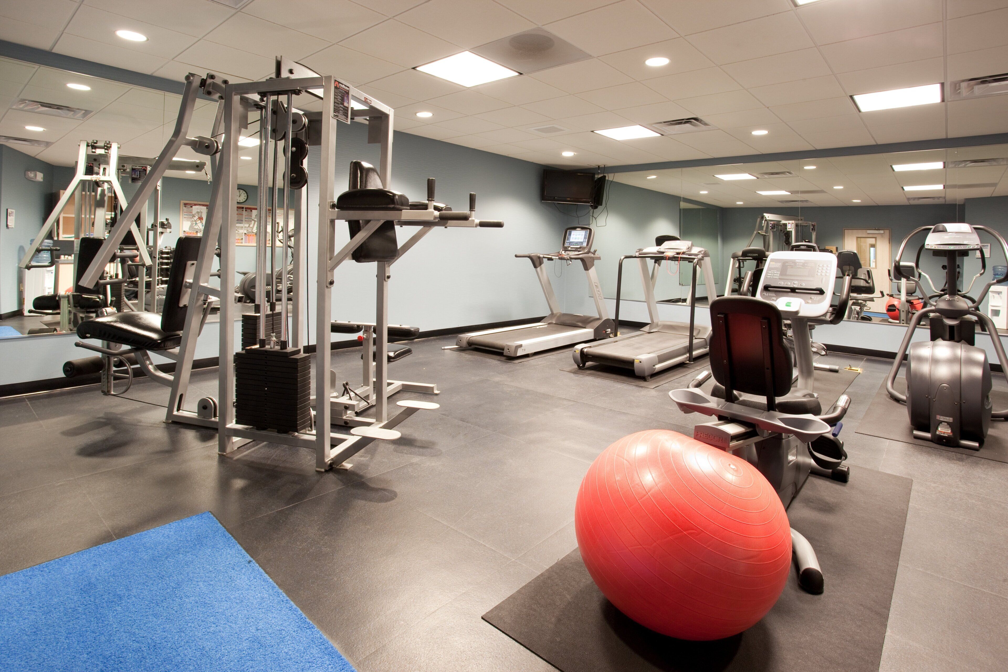 Ett gym med olika typer av träningsutrustning, inklusive löpband, en styrketräningsmaskin och en röd träningsboll.